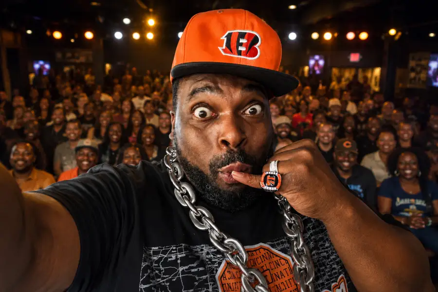 BIG Funny mugs wide-eyed for a selfie in a Cincinnati Bengals cap and chain, with a laughing crowd behind him.